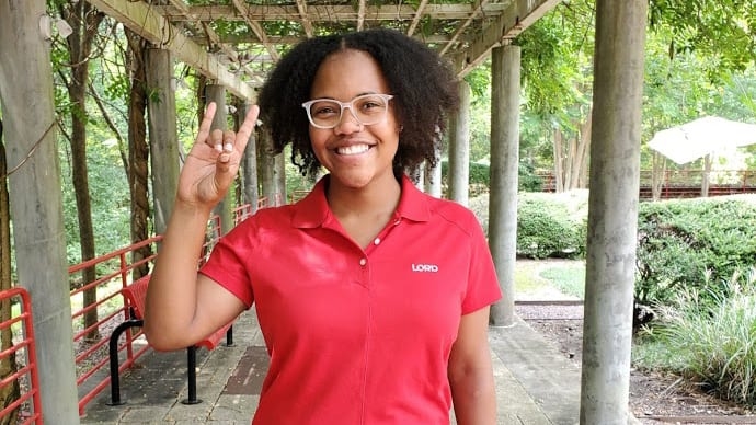 A female student makes the wolf ears sign while standing on a tree-lined sidewalk