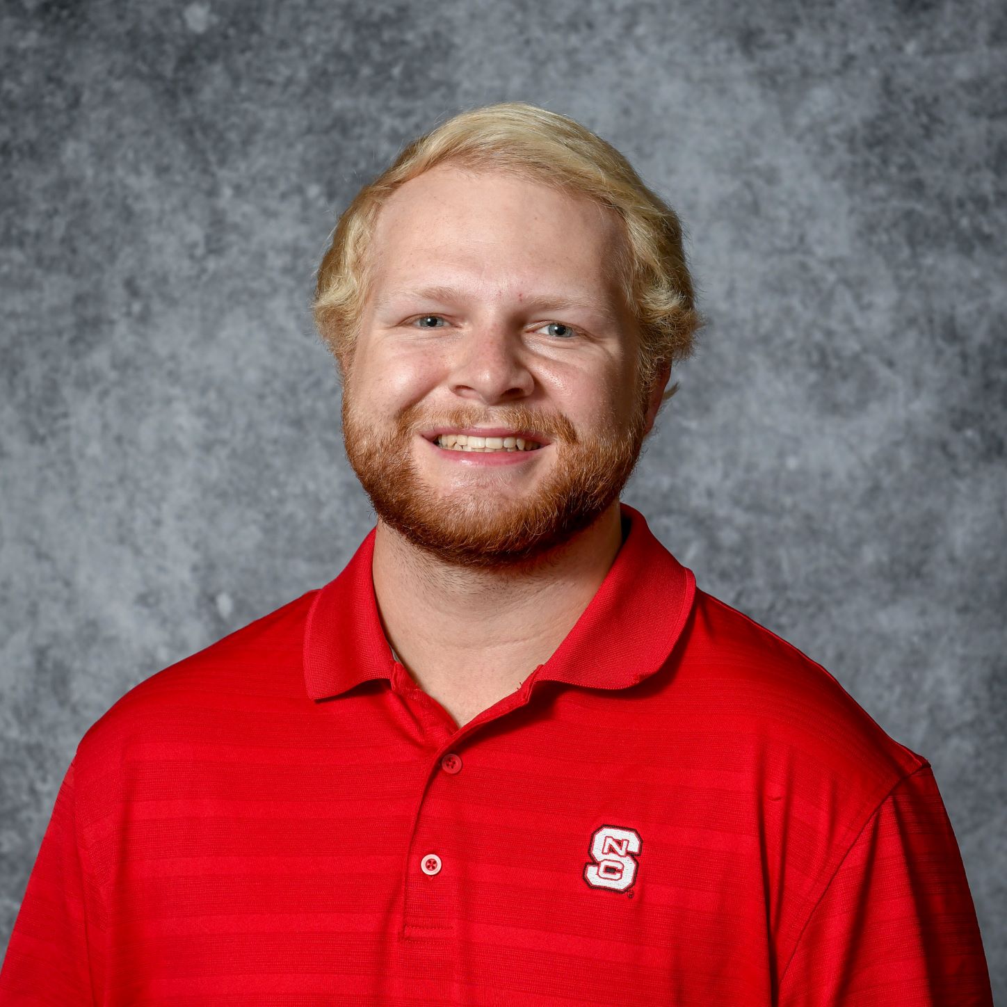The image represents the headshot of a young smiling adult with golden hair and a short beard wearing a red T-shirt before a grey background.