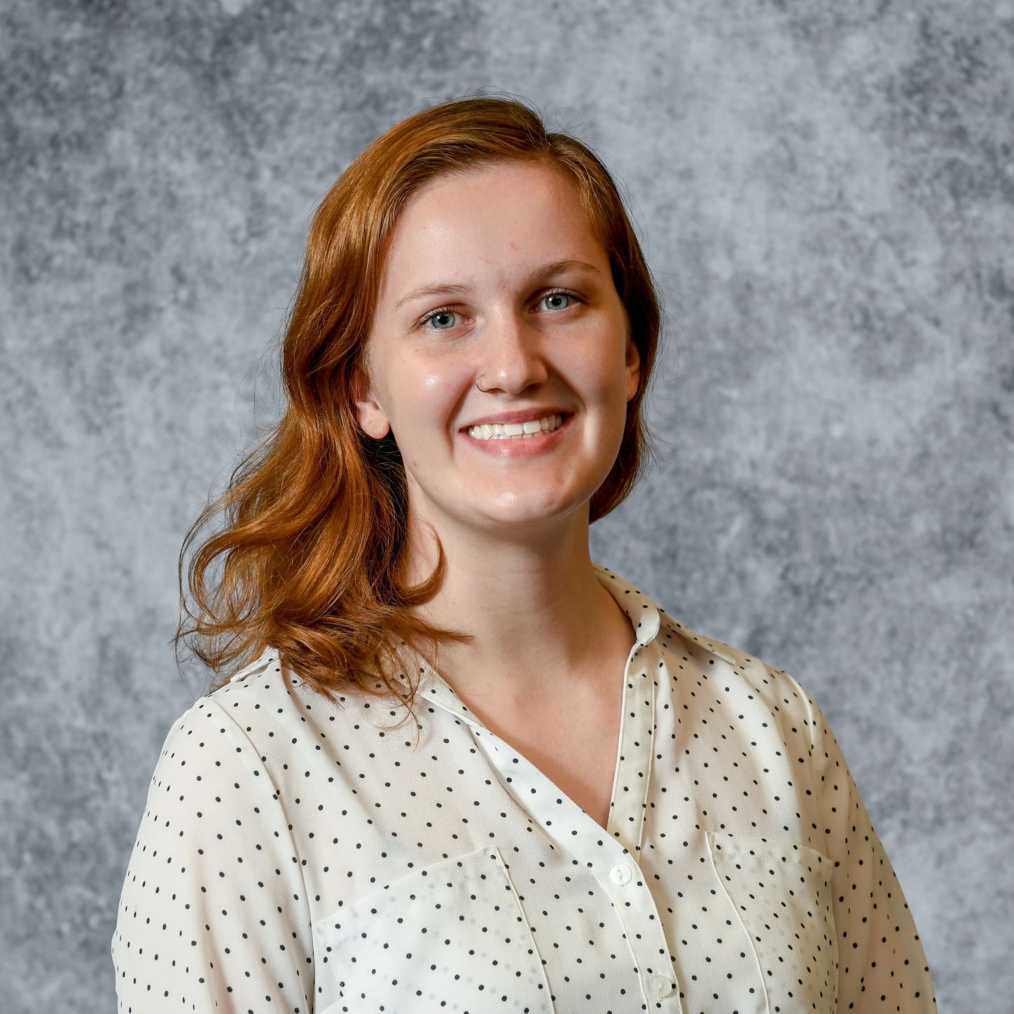 The image represents the headshot of graduate student Sarah Kromer wearing a printed button up shirt. The background behind is grey.