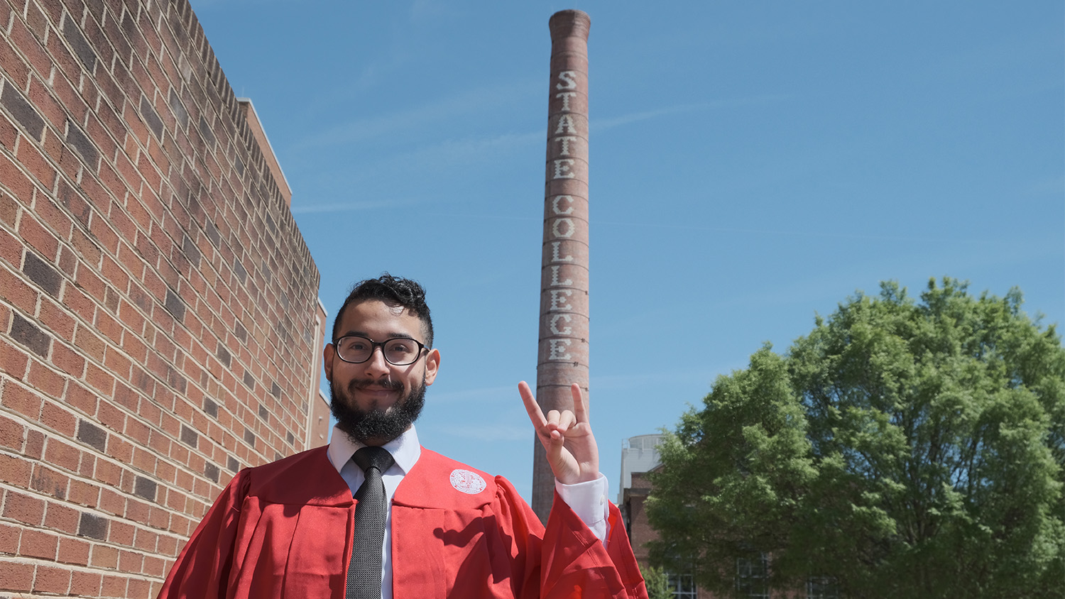 Arnulfo Silva wearing red graduation robes and making a wolfie sign with his hand in front of the smoke stack that says "State College."