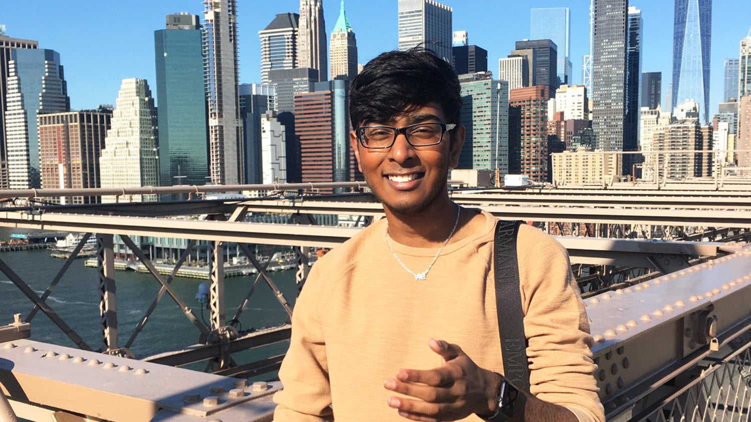 Kiran Soma is wearing eye glasses and a beige swetaer. He is posing in front of a bridge in a city crowded with high rises.
