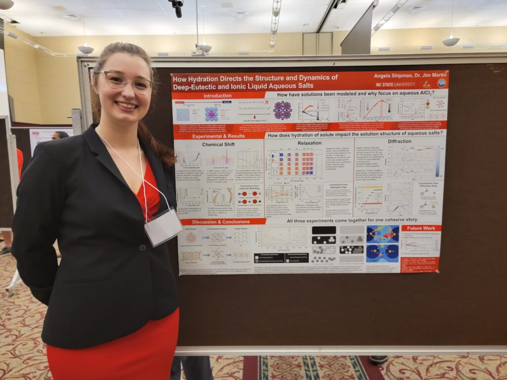 A young lady wearing red and black is standing by a research poster